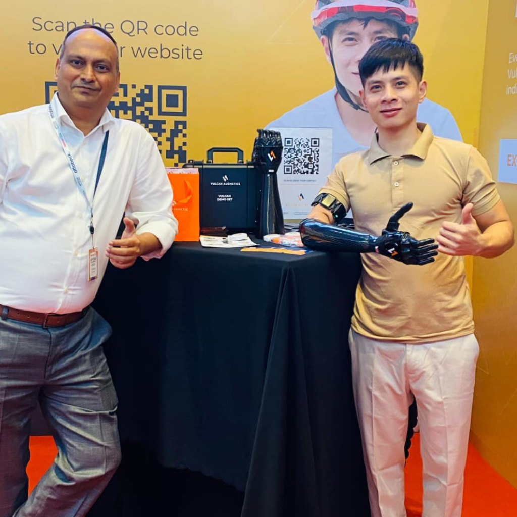 One young man wearing a black Vulcan prosthetic hand is taking a picture with another Indian man who is wearing a white shirt in an event hall