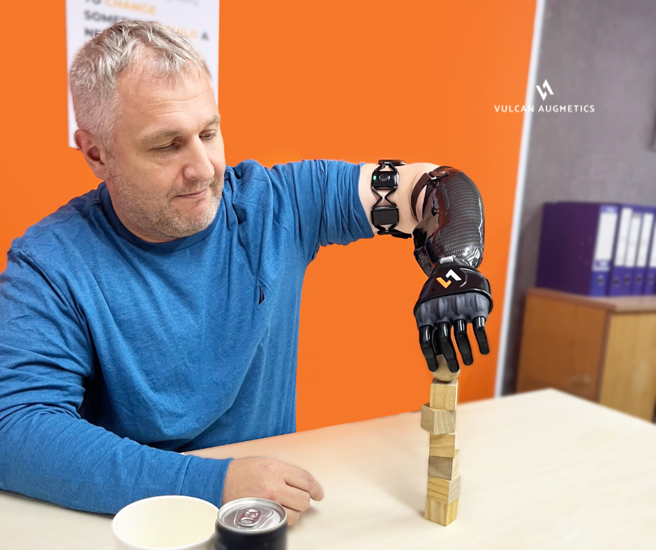 A male arm amputee wears a black Vulcan prosthetic hand and uses it to stack wooden cube in an orange room