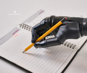 A young male arm amputee wears a black Vulcan prosthetic hand to hold a pencil and write note on a notebook