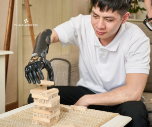 A young arm amputee man wears a black Vulcan prosthetic hand to play jenga with his friend in a brown living room